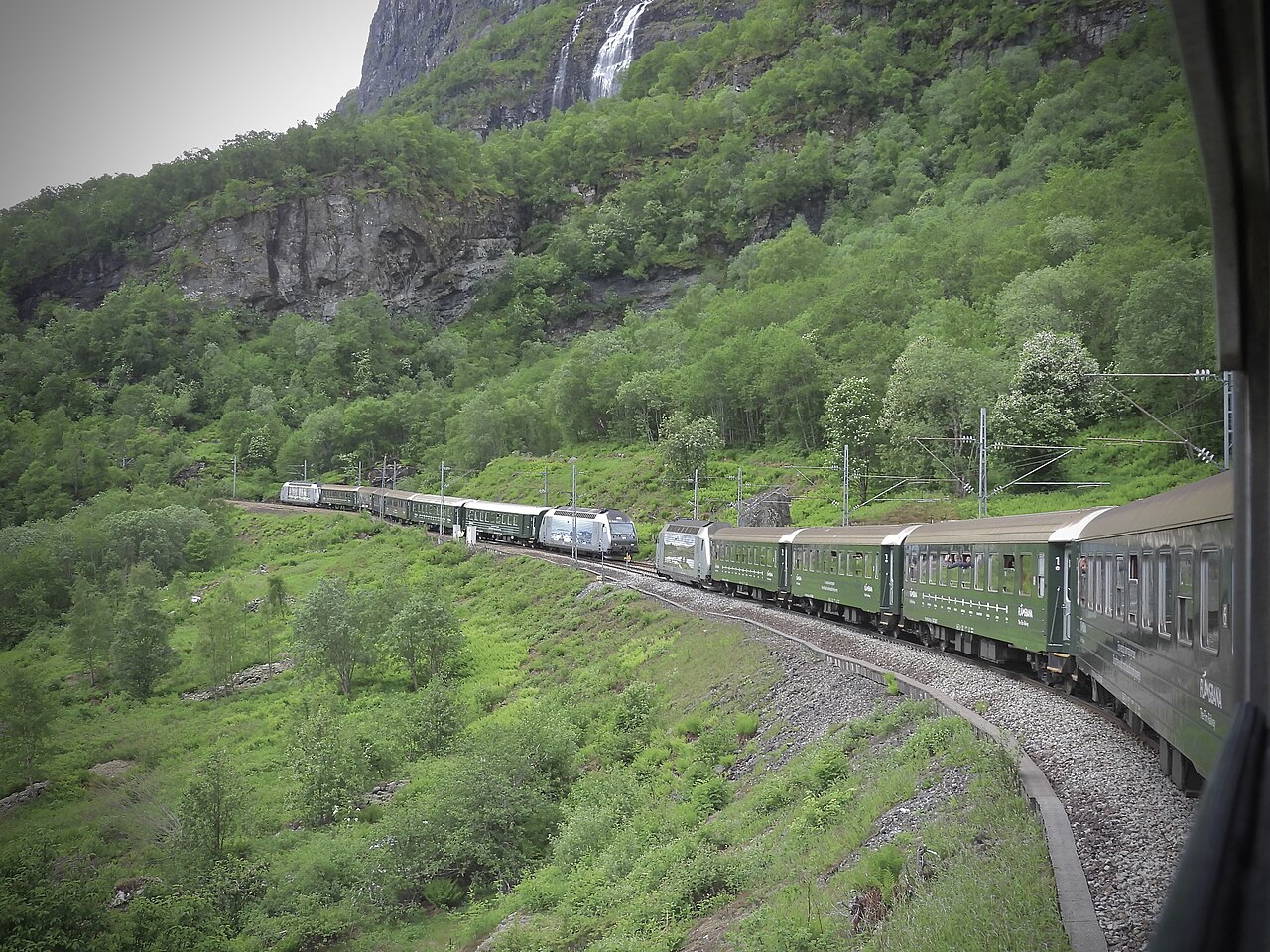 Green Flåm Railway trains passing beneath a waterfall in a steep Norwegian valley