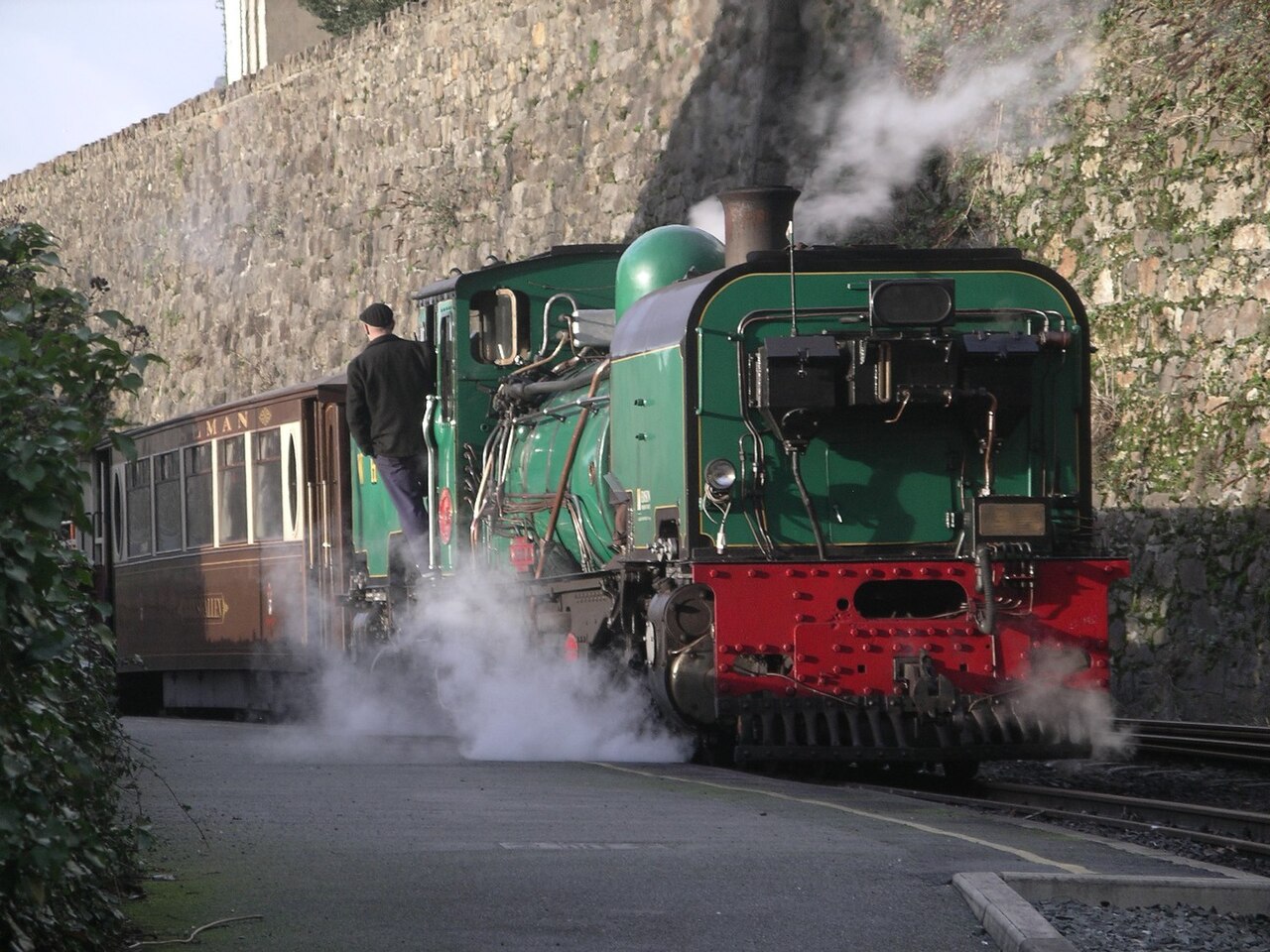 Green Welsh Highland steam locomotive and heritage cars beside a stone wall