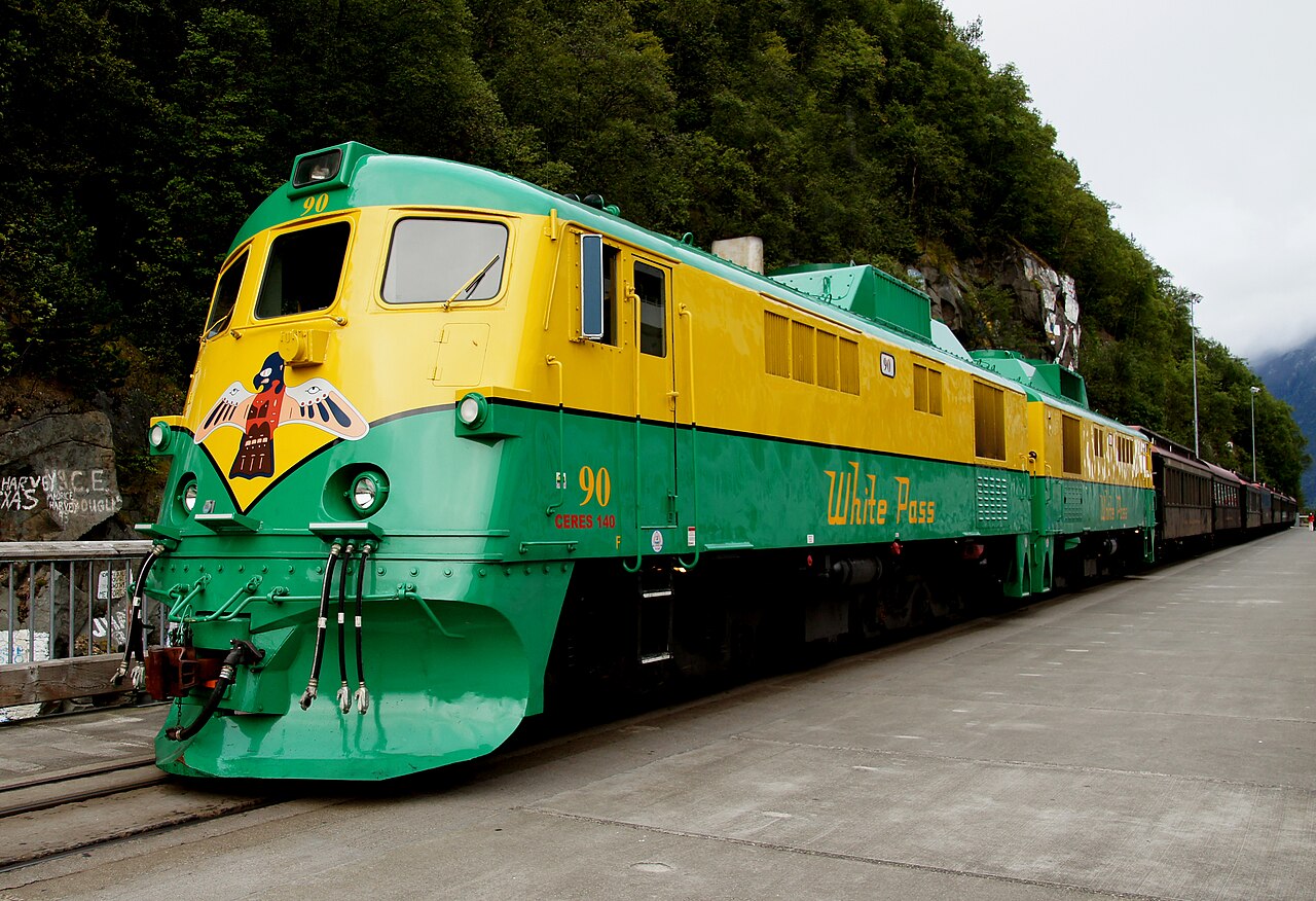 Green-and-yellow White Pass locomotive and passenger cars beside a rocky hillside